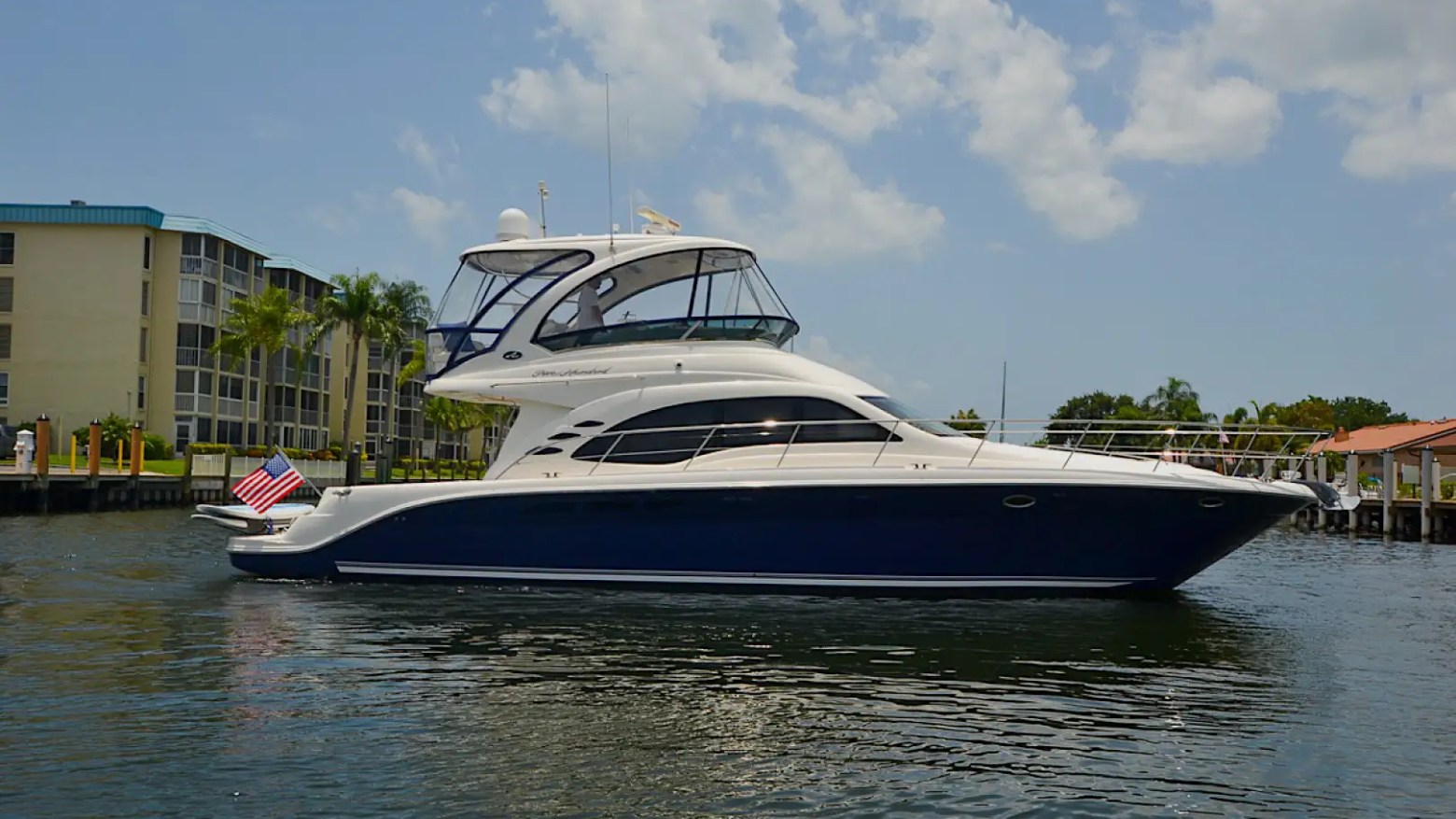Luxury yacht with flag docked by waterfront apartments under a partly cloudy sky.