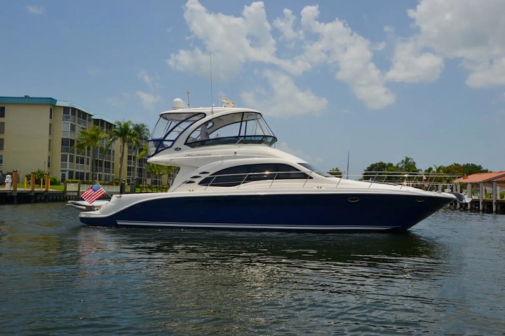 Luxury yacht with flag docked by waterfront apartments under a partly cloudy sky.