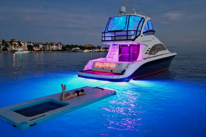 Yacht with pink lights anchored, a person on floating mat, glowing blue water at dusk.