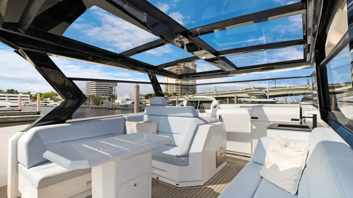 Modern yacht interior with sleek seating and glass roof, docked in a marina with city buildings visible.