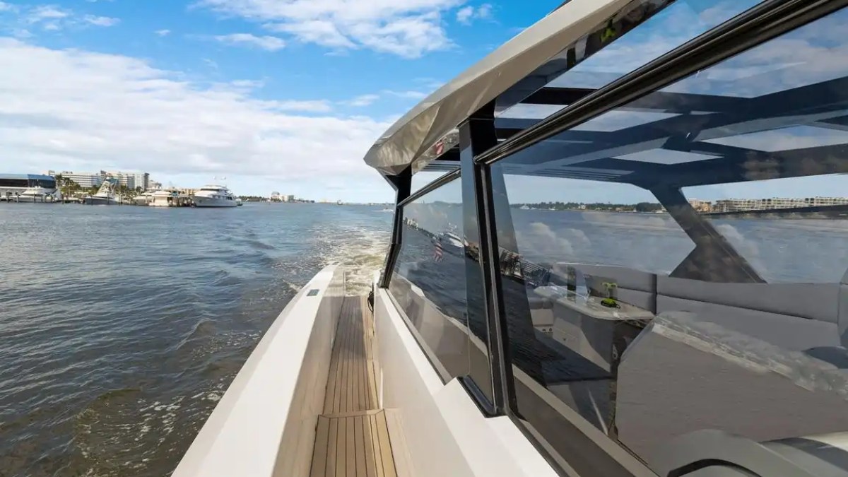 Side view of a yacht cruising on a wide river, with cityscape in the background, under a partly cloudy sky.