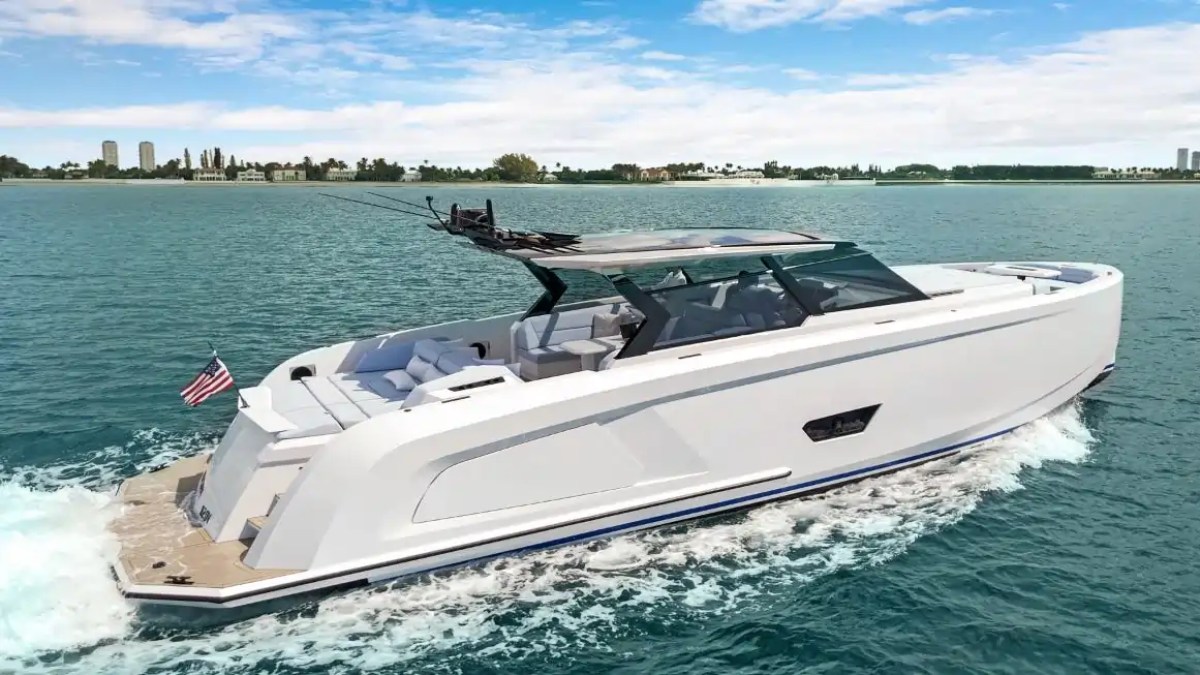 White yacht with American flag sailing on blue water, city skyline in background.