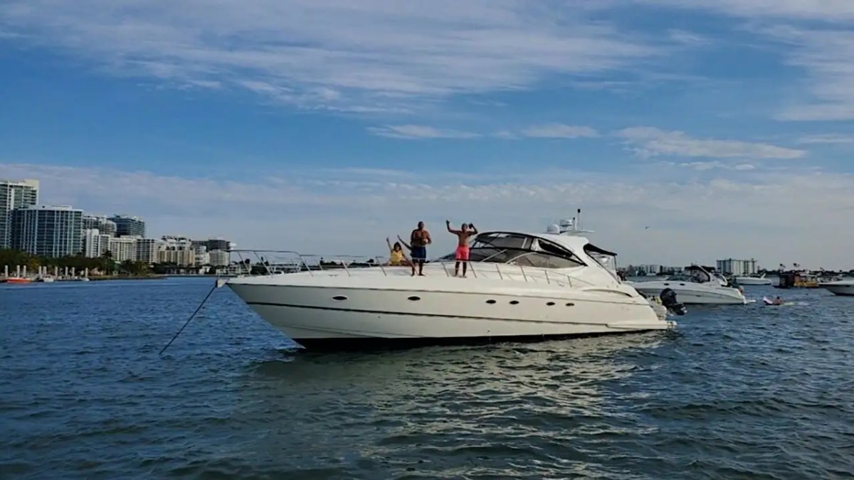 Three people on a yacht in a city harbor with buildings and a blue sky.