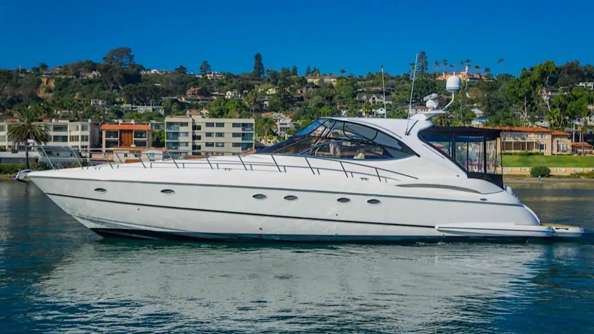 White yacht on calm water with a hillside town in the background under a clear blue sky.