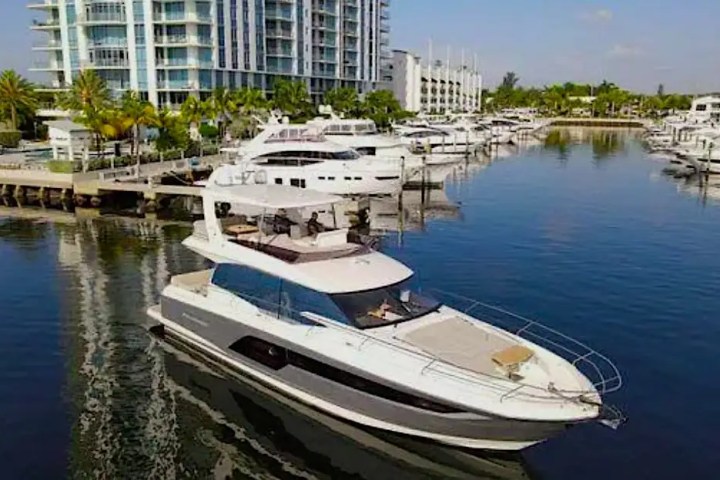 Modern yacht on calm water near marina with luxury apartments and boats in the background.