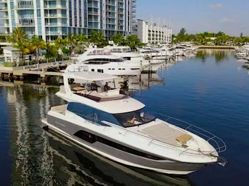 Modern yacht on calm water near marina with luxury apartments and boats in the background.
