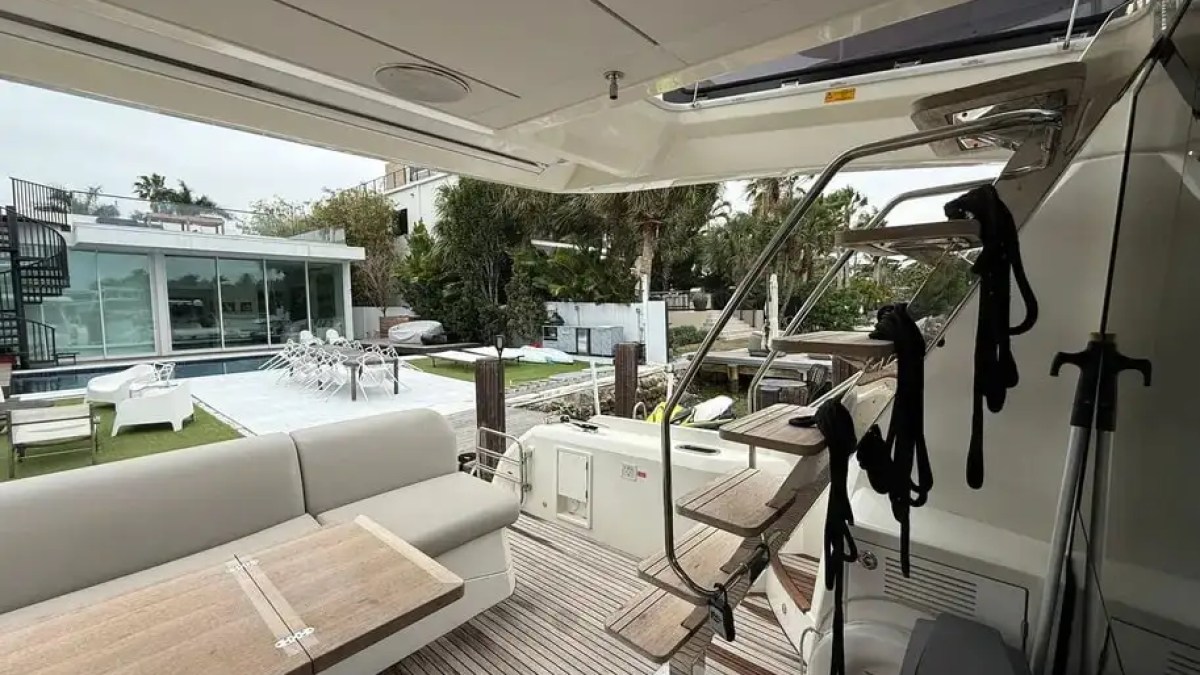View from a yacht's deck with seating, facing a modern house and pool in the background.