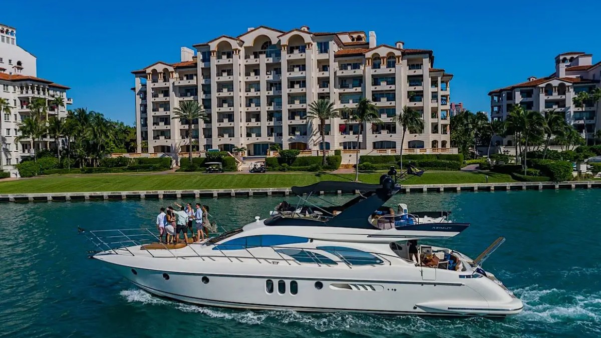 A yacht with people onboard cruising near luxury condos and palm trees.