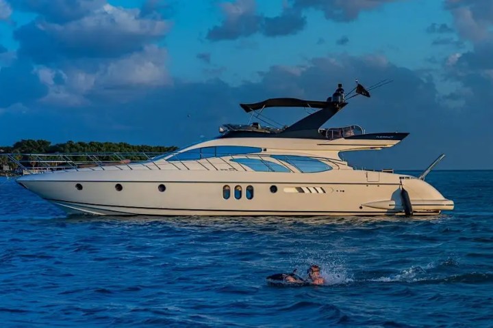 Luxury yacht on blue ocean water, with a person swimming nearby, under a cloudy sky.