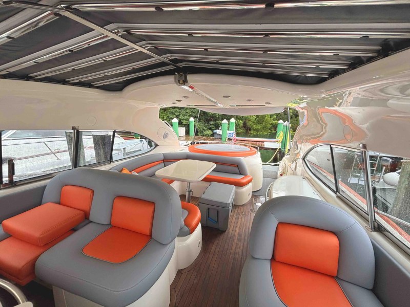 Interior of a boat with gray and orange seating, round table, and open views of a dock and greenery.