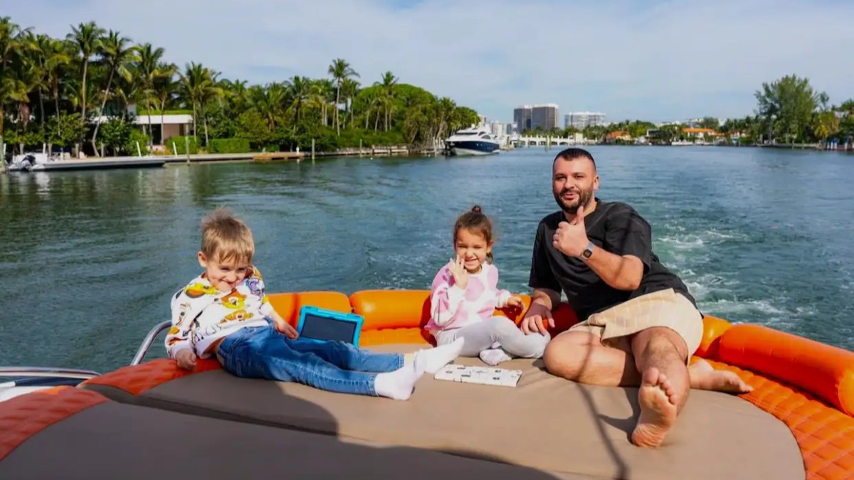Man with two children on a boat, smiling and giving thumbs up near a waterway with palm trees.