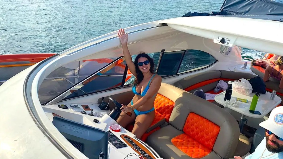 Person in blue bikini steering a boat, waving with one hand, in a sunlit cabin with orange seats.