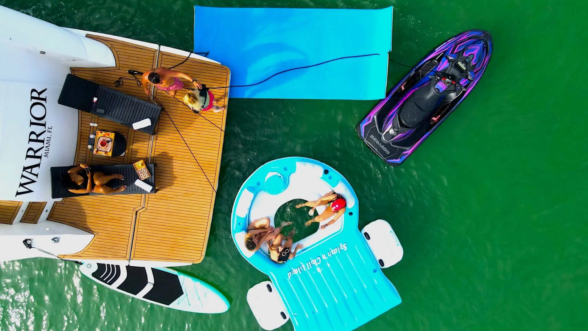 Aerial view of people on a yacht deck with paddleboard, pool float, and jet ski in green water.