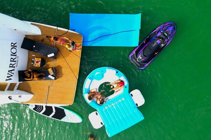 Aerial view of people on a yacht deck with paddleboard, pool float, and jet ski in green water.
