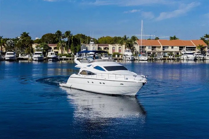 White yacht on a calm blue waterway with houses and trees in the background.