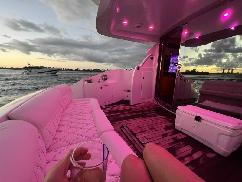 Boat interior with pink lighting, person holding drink, view of ocean and another boat at sunset.