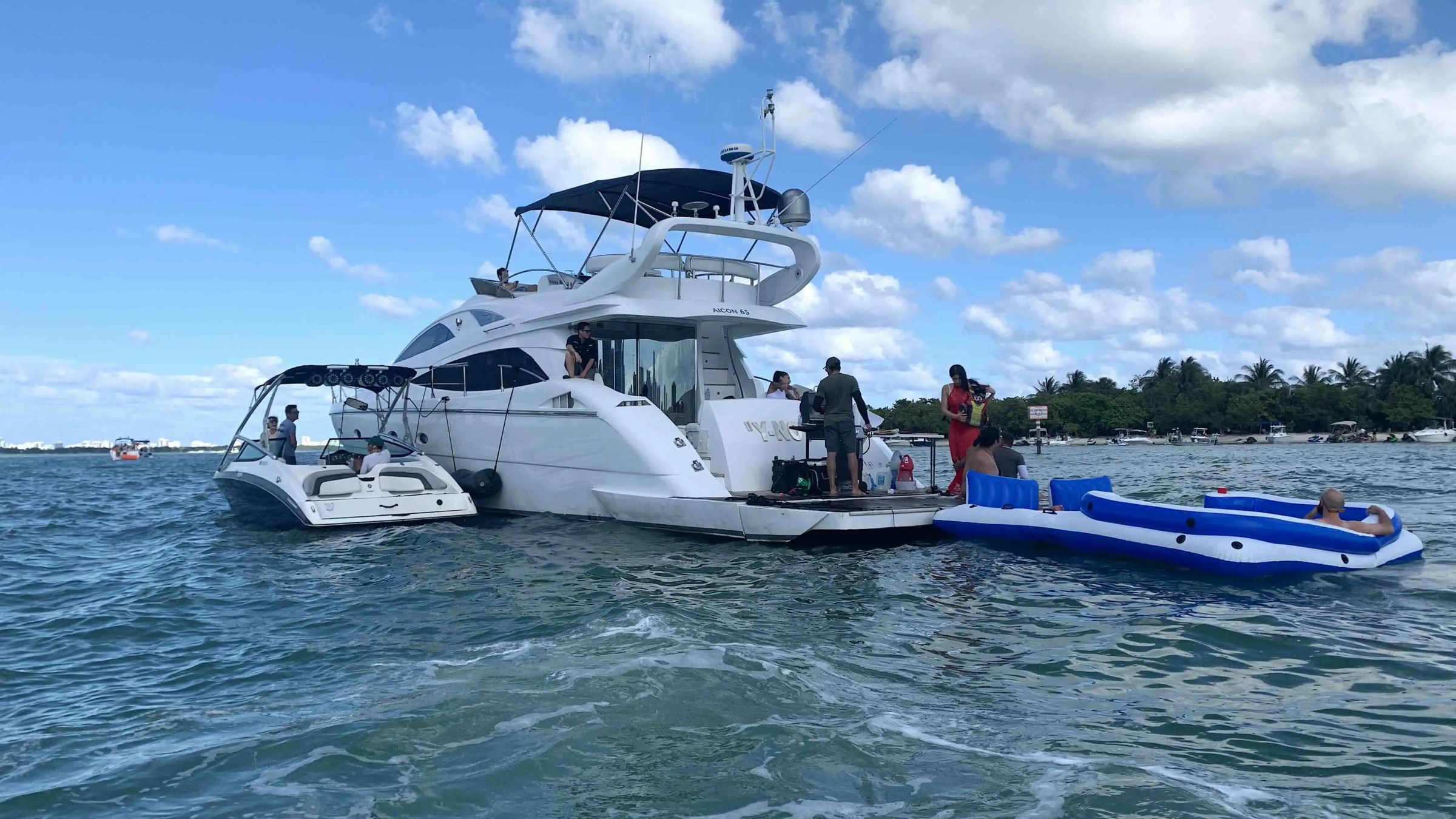 Two boats on the water with people and an inflatable raft nearby under a blue sky.