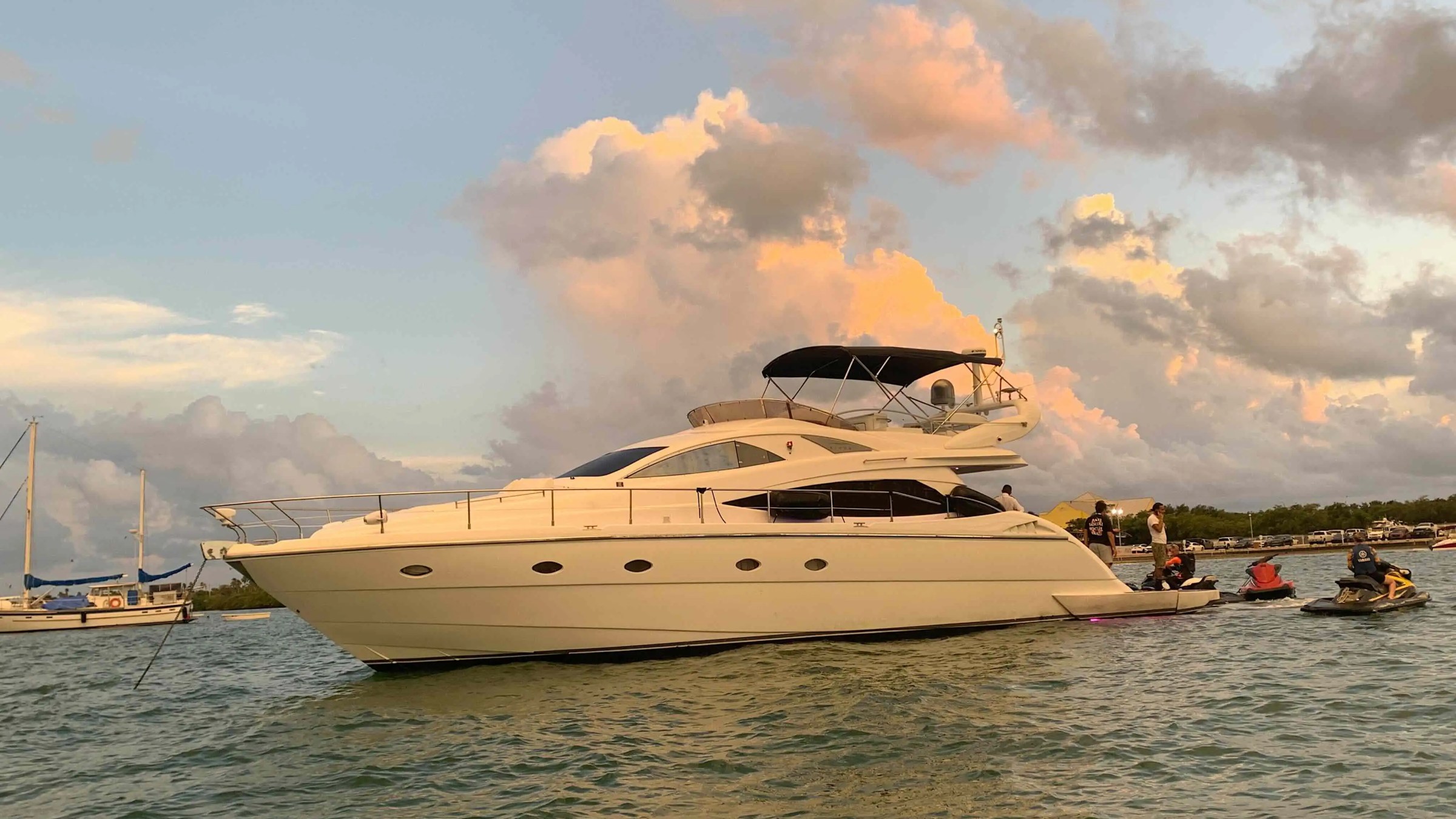 Luxury yacht on water during sunset with people on jet skis nearby.