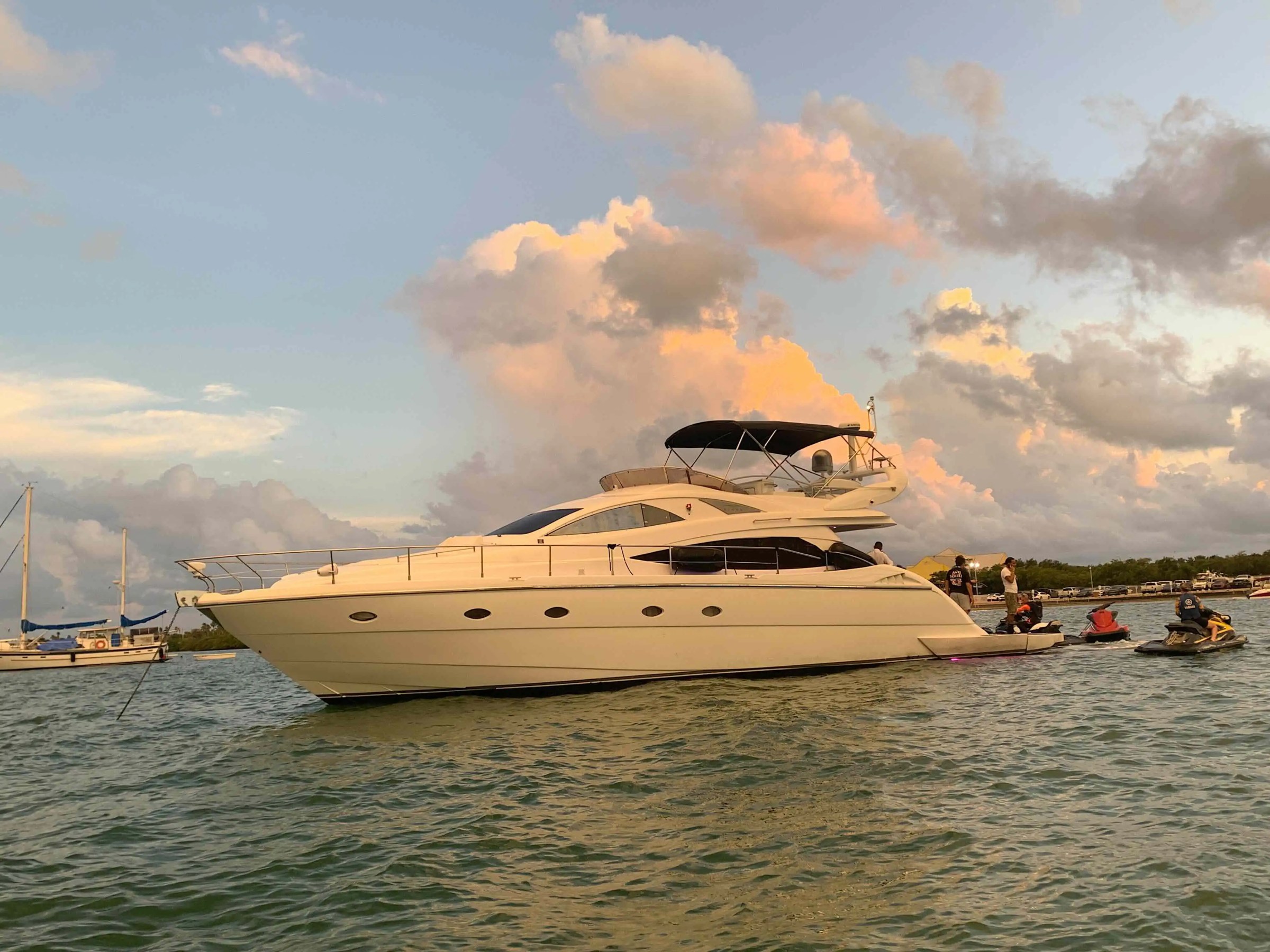 Luxury yacht on water during sunset with people on jet skis nearby.