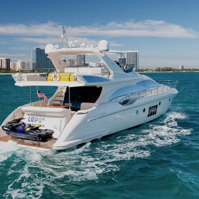 Luxury yacht with jet ski on deck sailing near city skyline in clear blue waters.