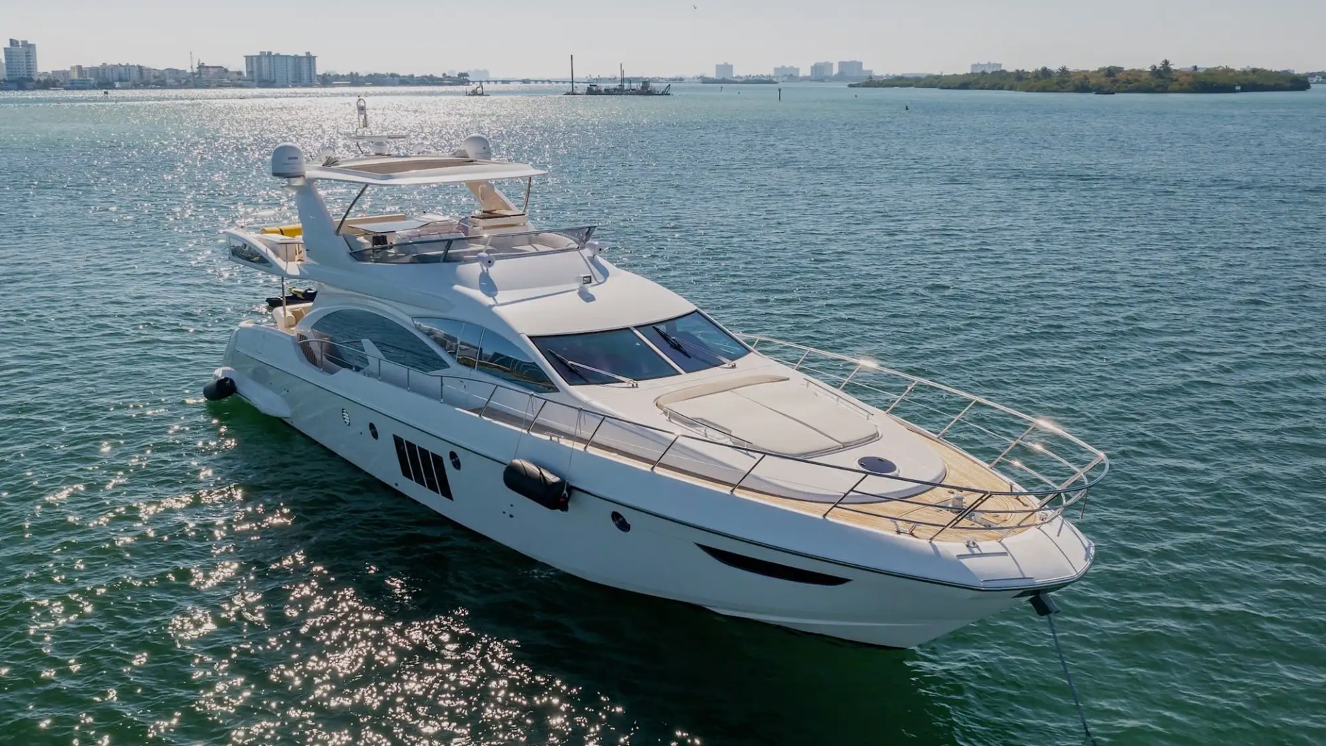 Luxury yacht anchored on a sunny day with city skyline and trees in the background.