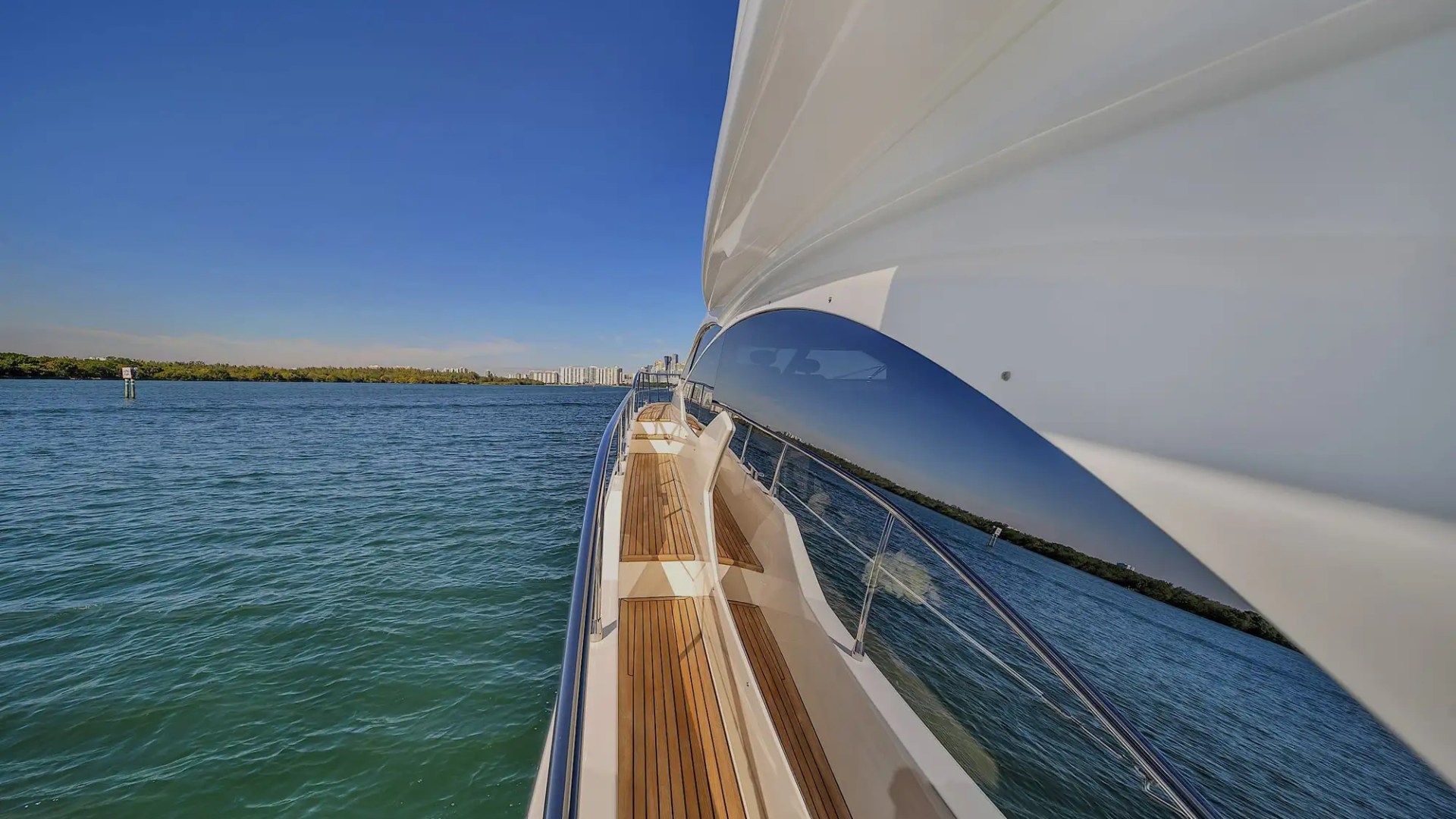 View along a yacht's side with wooden deck, railing, and water on a sunny day.