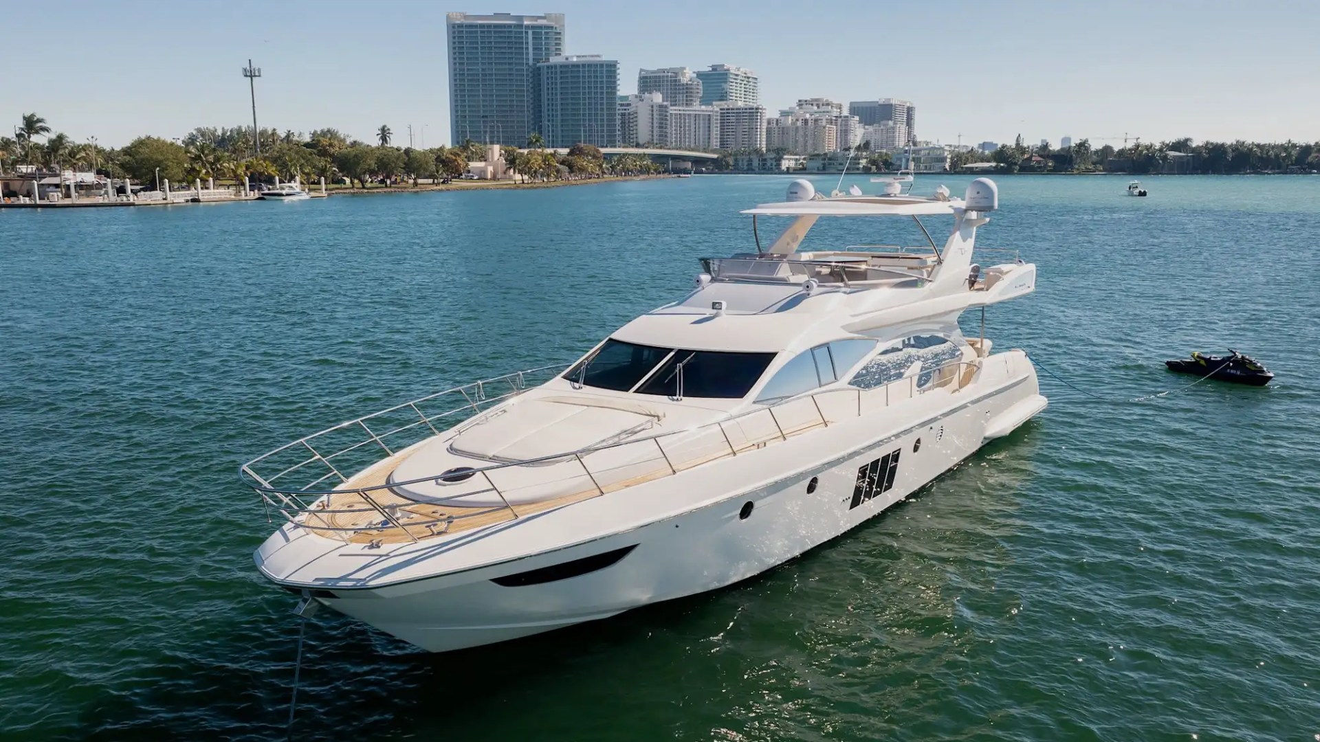 Luxury yacht on calm water with city skyline and trees in the background.