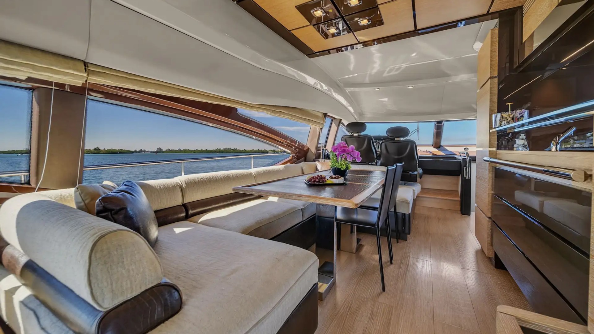 Luxurious yacht interior with wooden floors, plush seating, dining table, and scenic view through large windows.