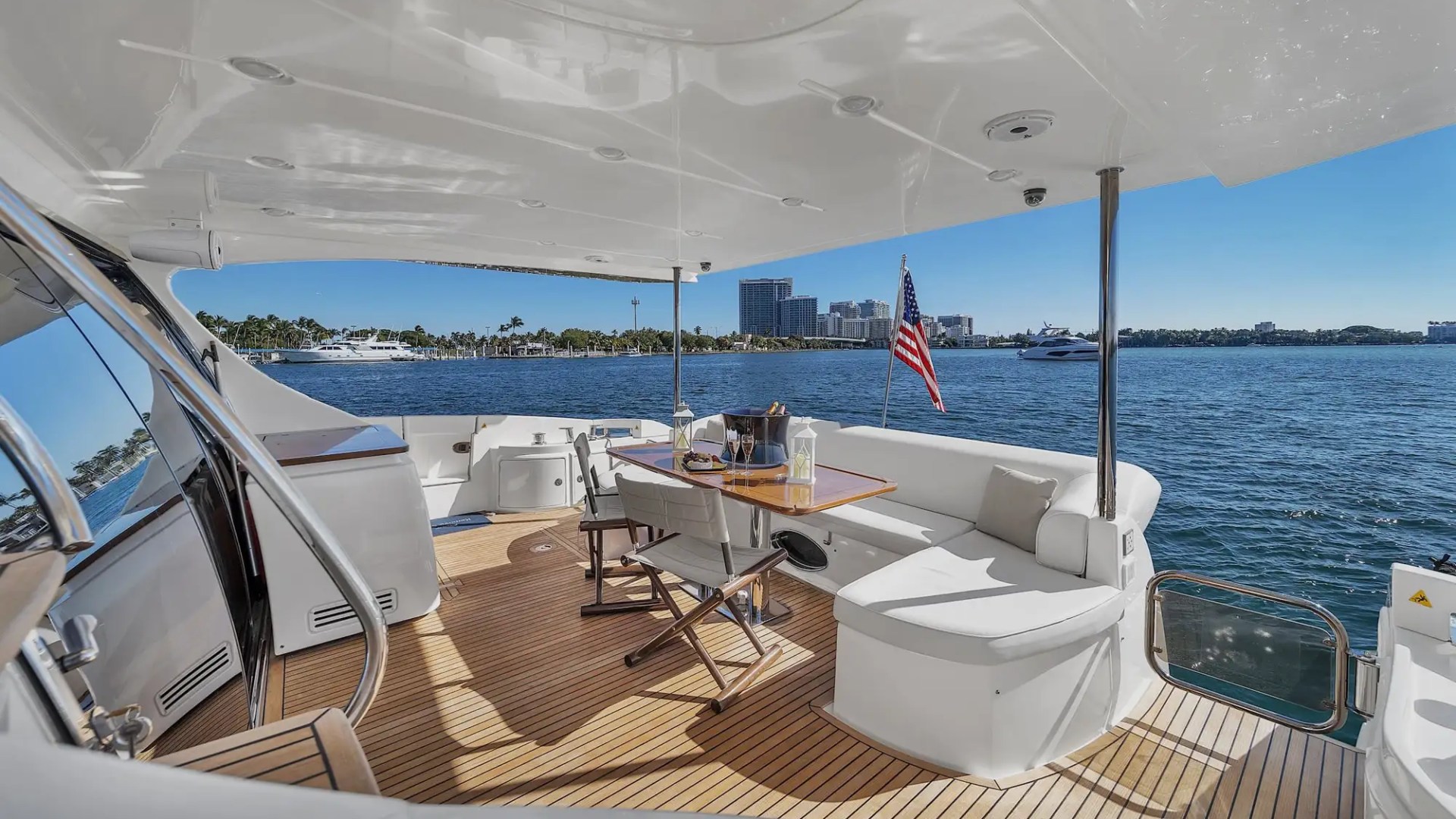 Yacht deck with seating, table, and American flag, overlooking city skyline and water.
