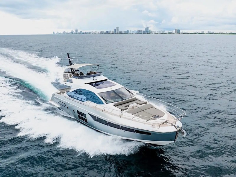 A yacht cruising on the ocean with a city skyline in the background under a cloudy sky.