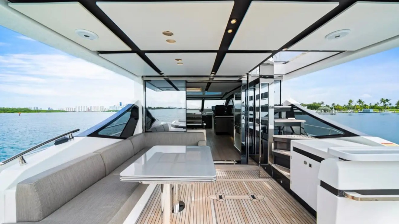 Modern yacht deck with seating area, table, and stairs, overlooking calm water and distant city skyline.