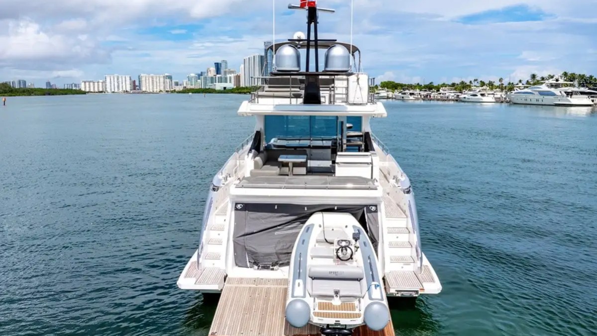 Large yacht with dinghy at the stern, city skyline in background, on a sunny day.