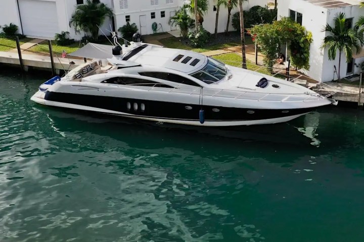 Sleek black and white yacht docked in a canal beside a house with palm trees.