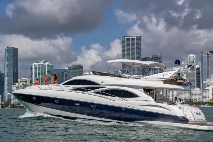 Luxury yacht cruising with city skyline in the background and people on deck.