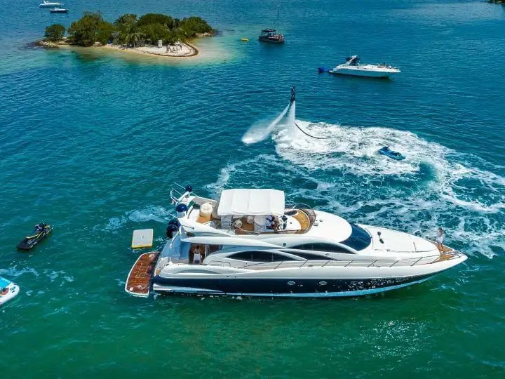 Luxury yacht near small island, with people on a raft and flyboarding in clear water.