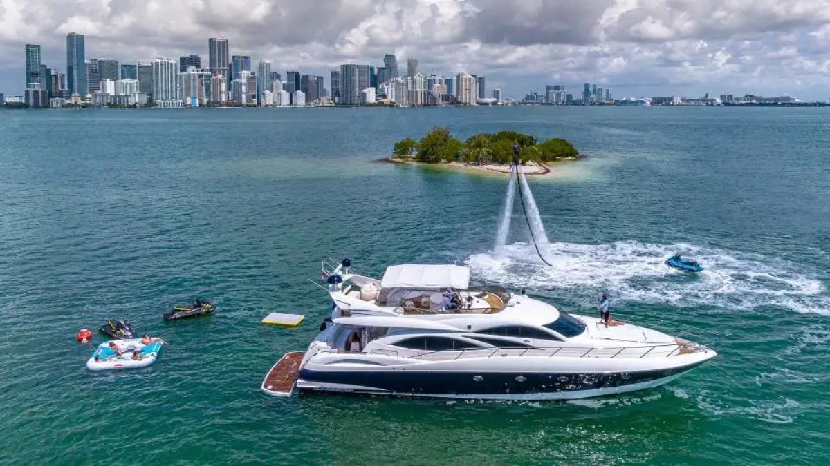 Luxury yacht on water with jet ski, island, and distant city skyline.