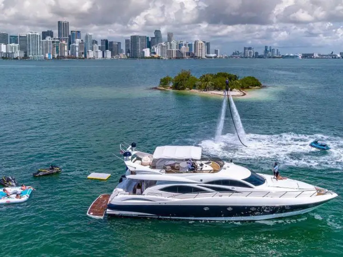 Luxury yacht on water with jet ski, island, and distant city skyline.