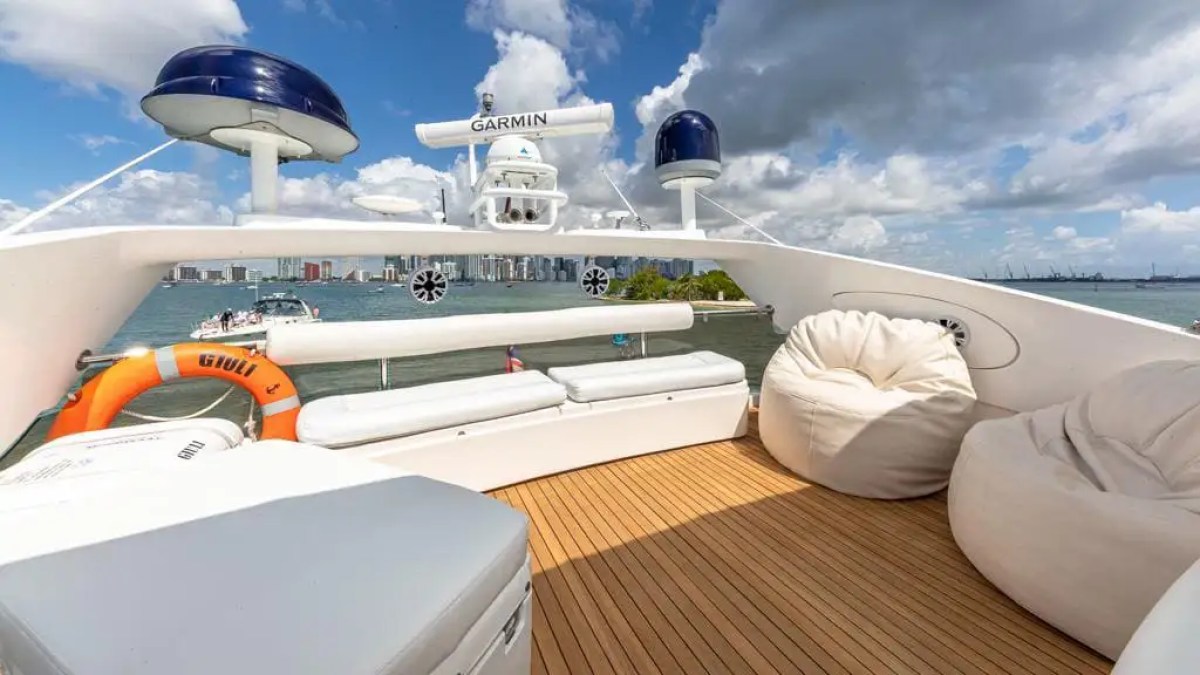Boat deck with white seating, bean bags, and a lifebuoy, overlooking a city skyline and cloudy sky.