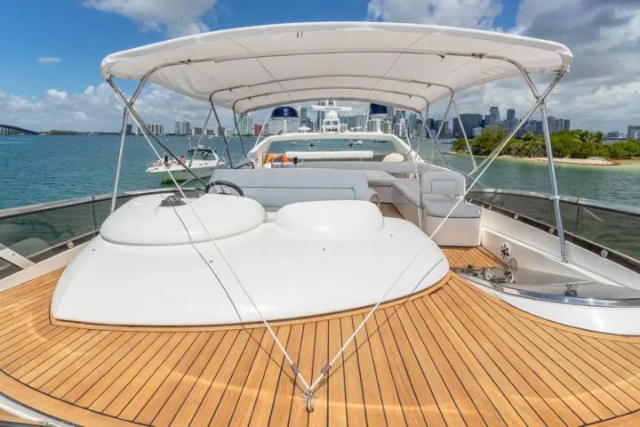 Luxury yacht deck with wooden flooring and canopy, city skyline and water in the background.