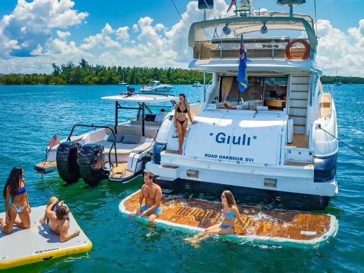 People on a yacht and floating mats in a sunny ocean setting.