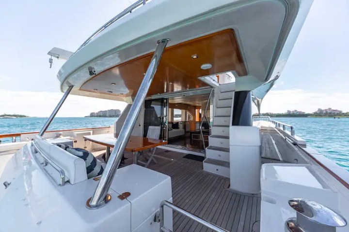 View of a yacht deck with seating, dining table, and stairs leading to upper levels, overlooking calm blue water.