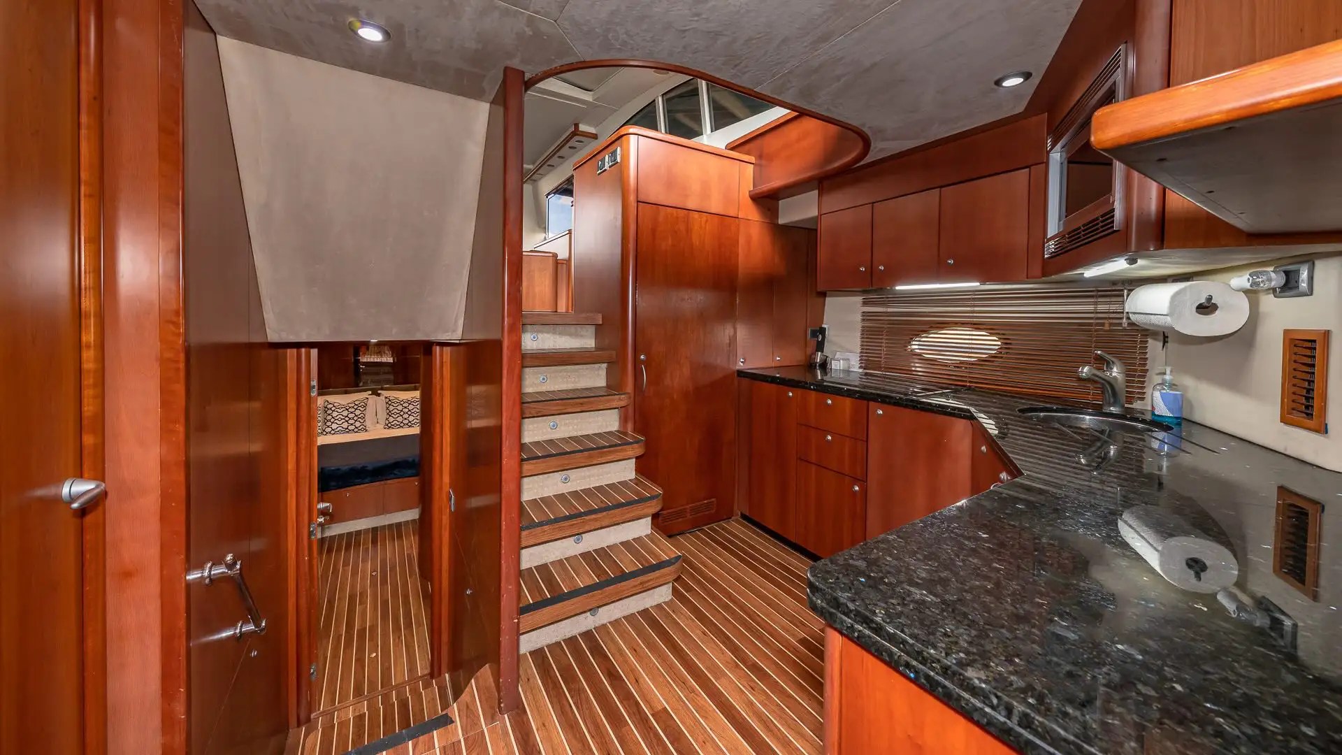 Wood-paneled yacht interior with stairs, kitchenette, and doorways, featuring dark countertops and modern lighting.