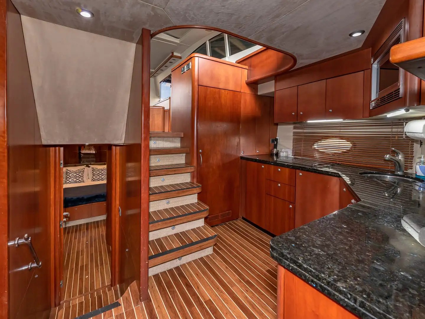 Wood-paneled yacht interior with stairs, kitchenette, and doorways, featuring dark countertops and modern lighting.