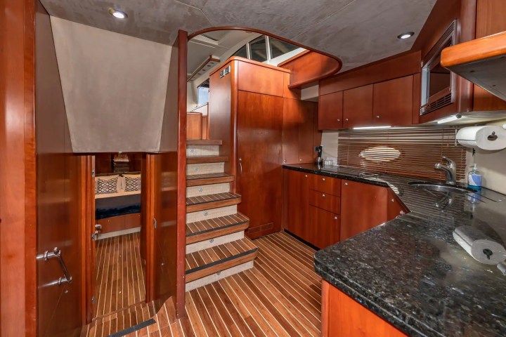 Wood-paneled yacht interior with stairs, kitchenette, and doorways, featuring dark countertops and modern lighting.