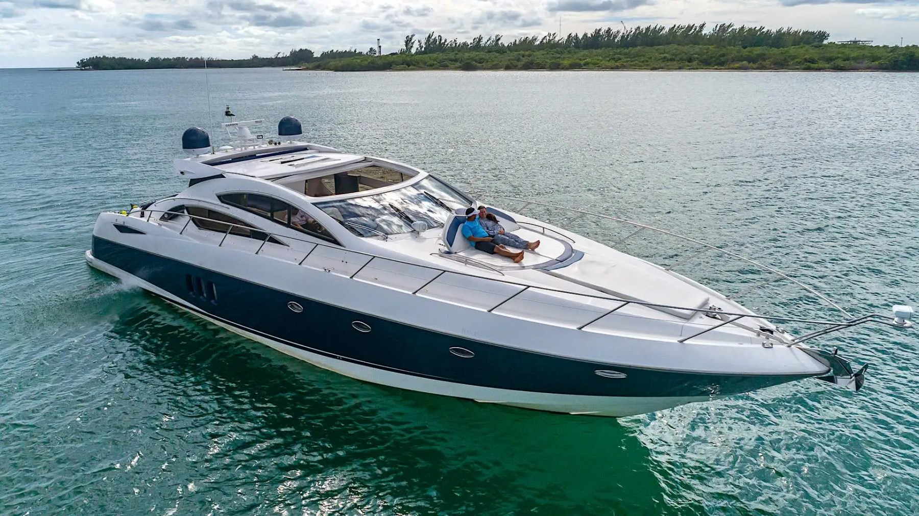 Luxury yacht on water with two people relaxing on the deck, cloudy sky and shoreline in background.