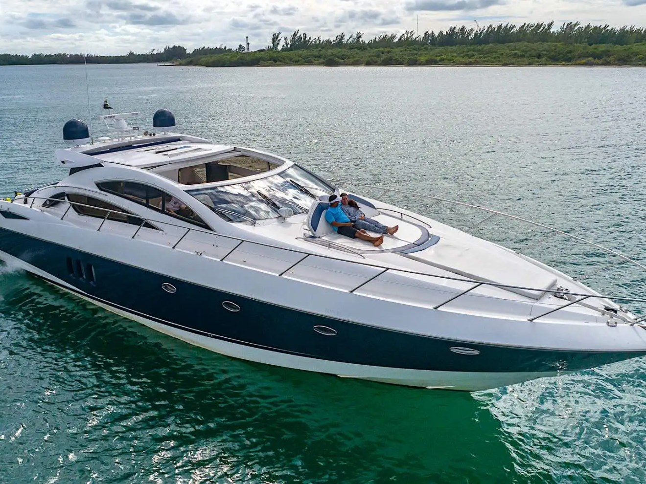 Luxury yacht on water with two people relaxing on the deck, cloudy sky and shoreline in background.