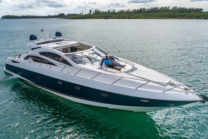 Luxury yacht on water with two people relaxing on the deck, cloudy sky and shoreline in background.