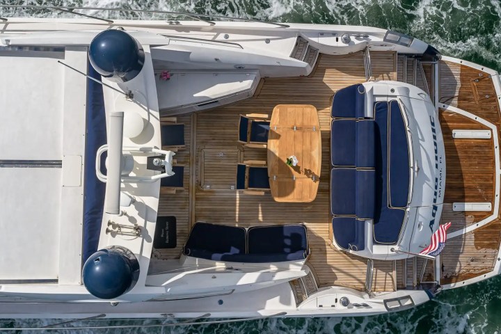 Aerial view of yacht with a wooden deck, seating, and table on the back, sailing through water.