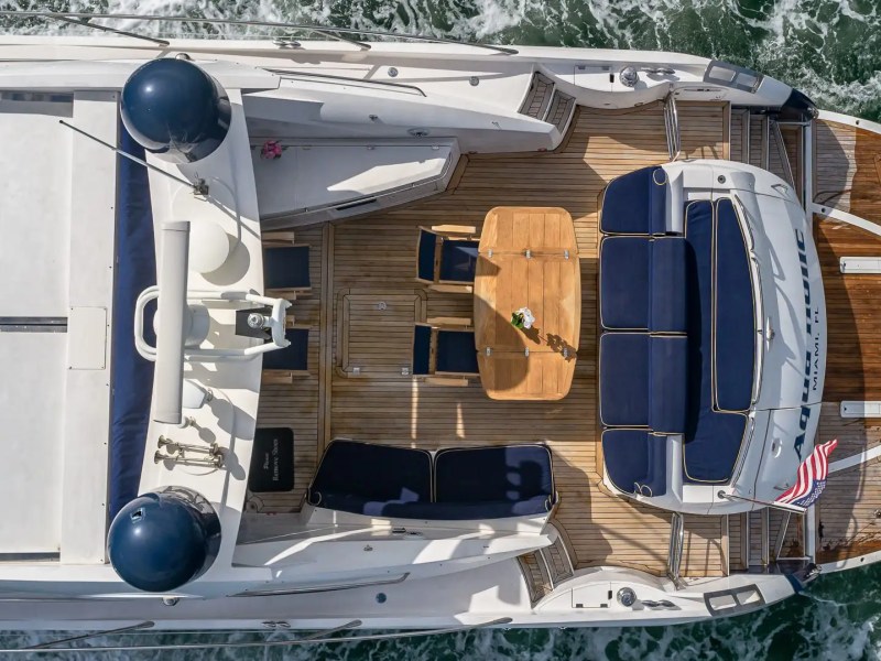 Aerial view of yacht with a wooden deck, seating, and table on the back, sailing through water.