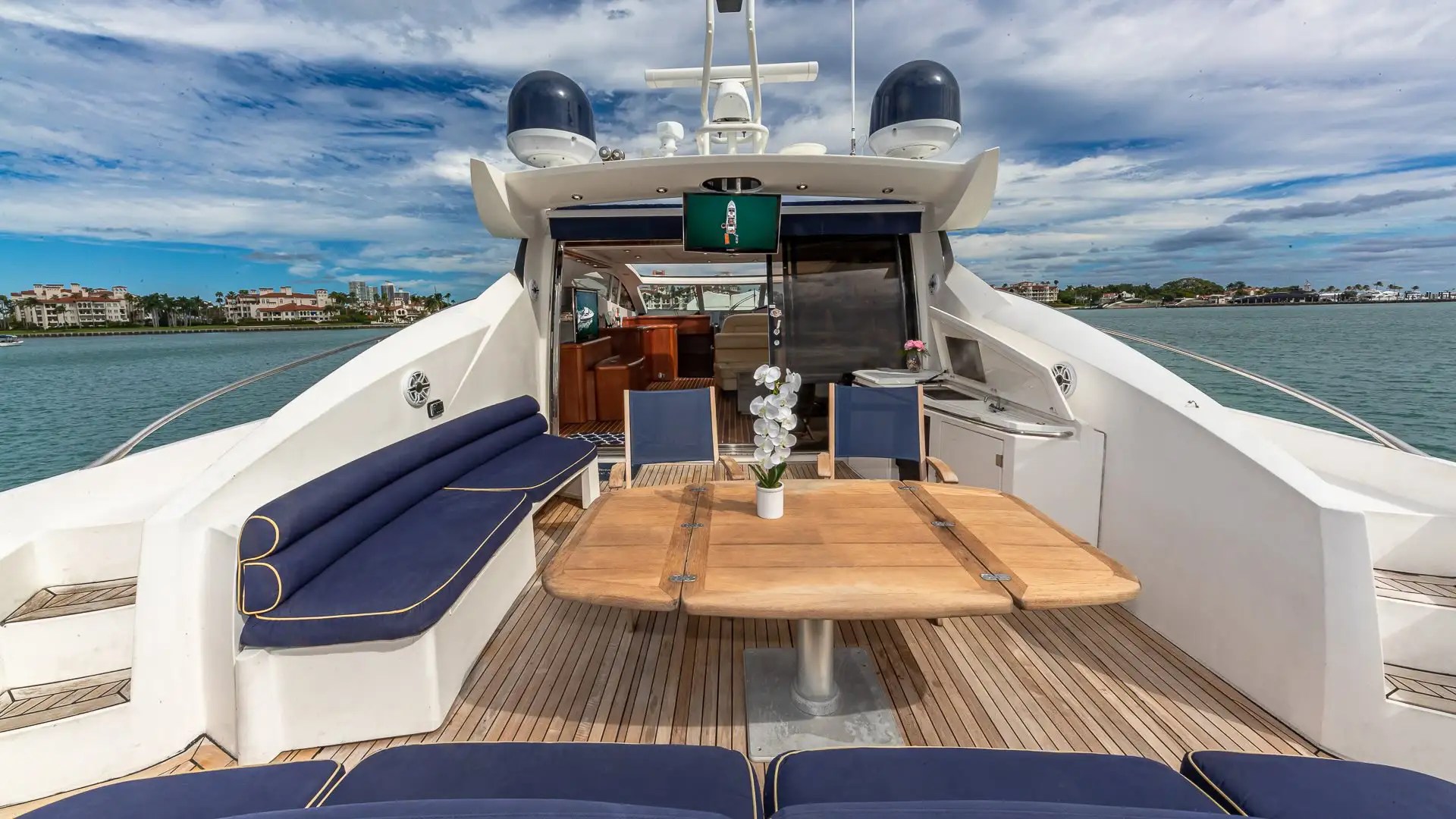 Luxury yacht deck with blue cushioned seating, wooden table, and view of buildings across the water.